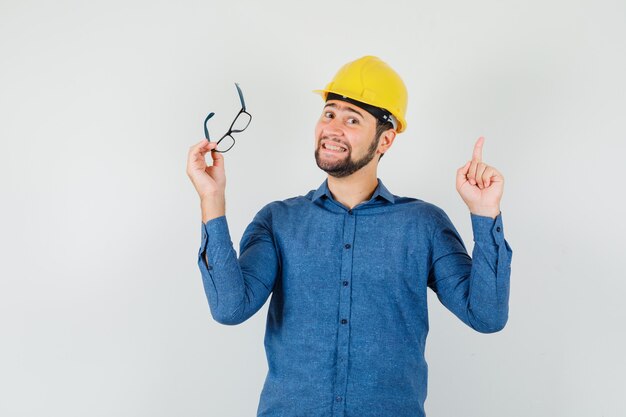 Jeune homme pointant vers le haut, tenant des lunettes en chemise, casque et regardant joyeux.