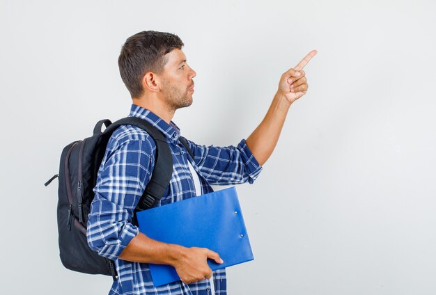 Jeune homme pointant vers le haut tout en tenant le presse-papiers en chemise, sac à dos et à la recherche concentrée.