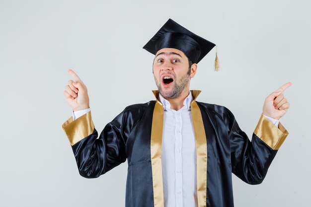 Jeune homme pointant vers le haut en uniforme d'études supérieures et à la recherche d'excité. vue de face.