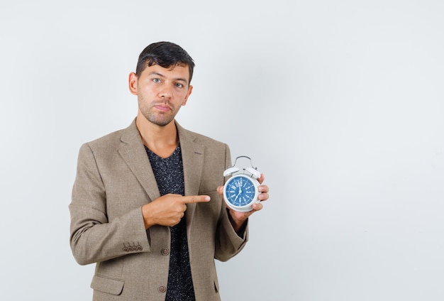 Jeune homme pointant vers l'horloge en veste marron grisâtre et regardant concentré, vue de face. espace pour le texte