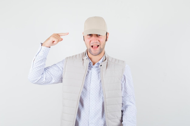 Jeune homme pointant vers sa casquette en chemise, veste sans manches, casquette et à l'optimiste, vue de face.