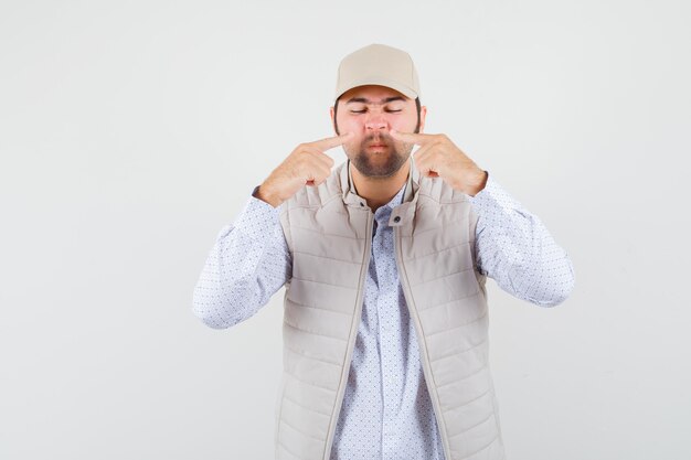 Jeune homme pointant vers son nez en chemise, veste sans manches, casquette et à la recherche concentrée. vue de face.