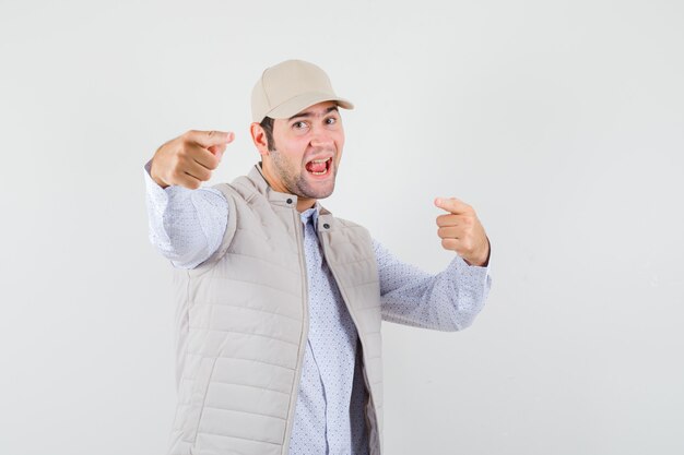 Jeune homme pointant en veste beige et casquette et à la recherche de plaisir. vue de face.