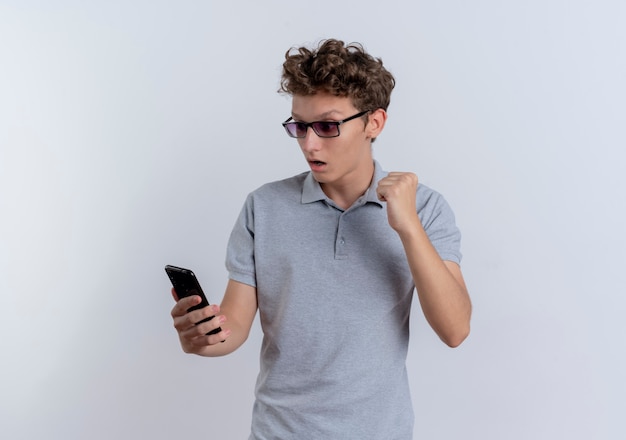 Jeune homme en polo gris regardant l'écran de son smartphone serrant le poing heureux et excité debout sur un mur blanc