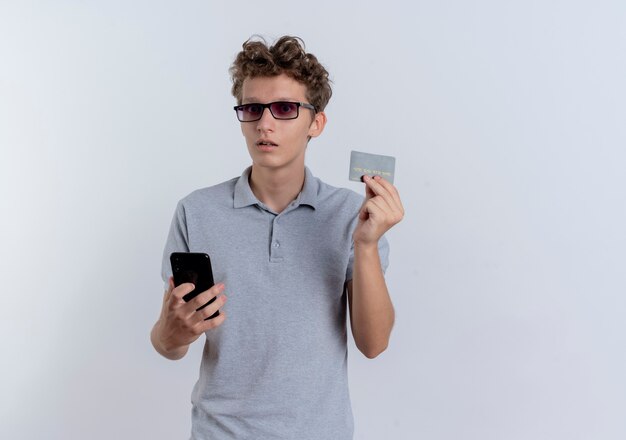 Jeune homme en polo gris tenant smartphone montrant la carte de crédit étant confus debout sur un mur blanc