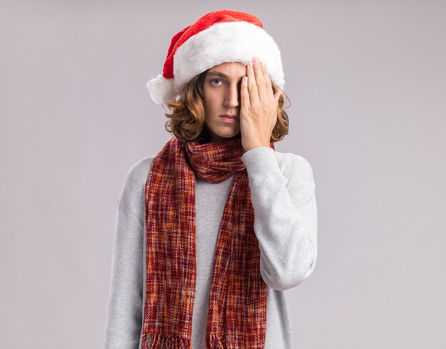 Jeune homme portant un bonnet de Noël avec écharpe chaude autour de son cou regardant la caméra avec un visage sérieux couvrant un œil avec la main debout sur fond blanc