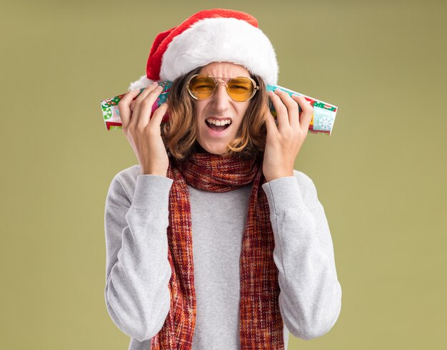 Jeune homme portant un bonnet de noel et des lunettes jaunes avec une écharpe chaude autour du cou couvrant ses oreilles avec des gobelets en papier colorés avec une expression agacée debout sur un mur vert