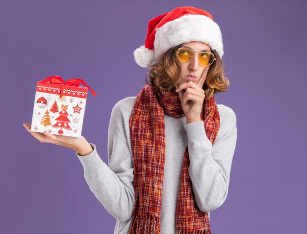 Jeune homme portant un bonnet de noel et des lunettes jaunes avec une écharpe chaude autour du cou tenant un cadeau de noël levant avec une expression pensive pensant debout sur un mur violet