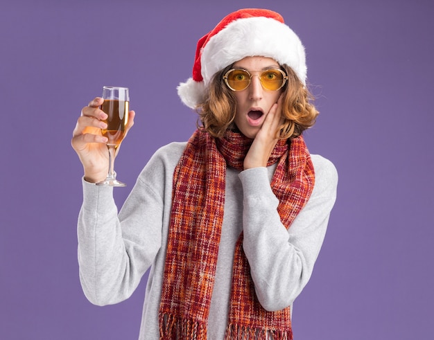 Jeune homme portant un bonnet de noel et des lunettes jaunes avec une écharpe chaude autour du cou tenant un verre de champagne étonné et surpris debout sur un mur violet