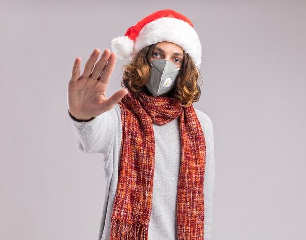 Jeune homme portant un bonnet de noel et un masque de protection faciale avec une écharpe chaude autour du cou avec un visage sérieux faisant un geste d'arrêt avec la main debout sur un mur blanc