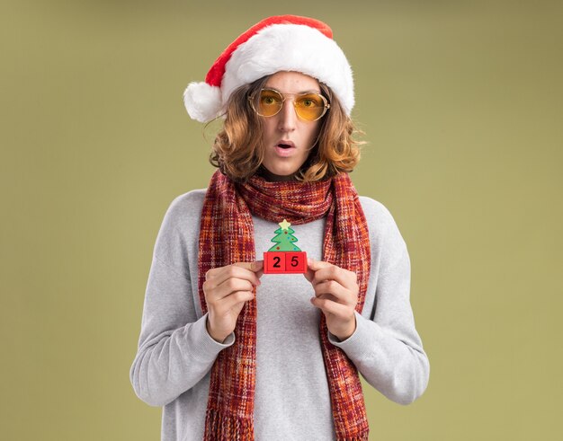 Jeune homme portant un bonnet de noel de Noël et des lunettes jaunes avec une écharpe chaude autour du cou tenant des cubes de jouet avec date vingt-cinq regardant la caméra confus et surpris debout sur fond vert