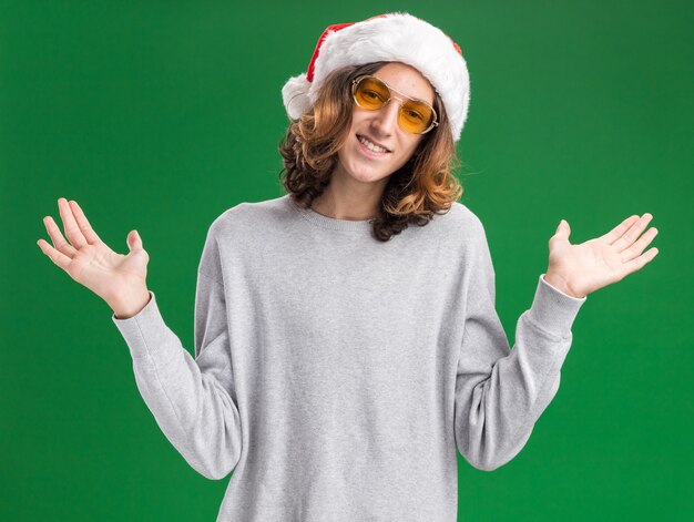 Jeune homme portant un bonnet de noel de Noël et des lunettes jaunes regardant la caméra avec le sourire sur le visage en écartant les bras sur les côtés debout sur fond vert