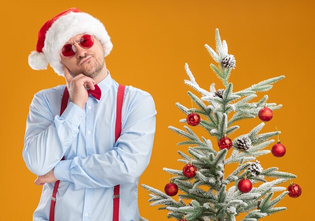 Jeune homme portant des bretelles noeud papillon en bonnet de Noel et lunettes rouges debout à côté de l'arbre de Noël avec une expression pensive sur le visage pensant au mur orange