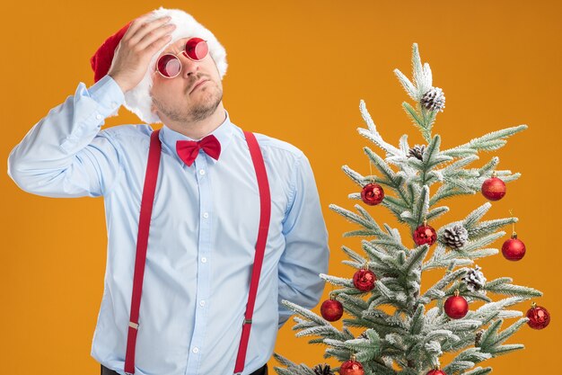 Jeune homme portant des bretelles noeud papillon en bonnet de Noel et lunettes rouges debout à côté de l'arbre de Noël à la perplexité avec la main sur la tête sur le mur orange