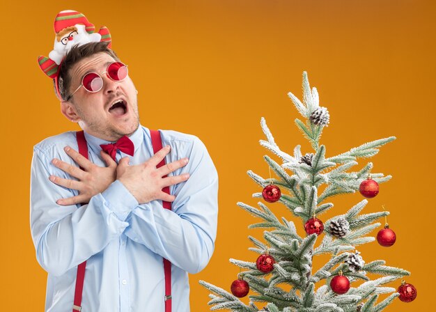 Jeune homme portant des bretelles noeud papillon dans la jante avec le père Noël et des lunettes rouges debout à côté de l'arbre de Noël en regardant étonné et inquiet se tenant la main sur sa poitrine sur fond orange