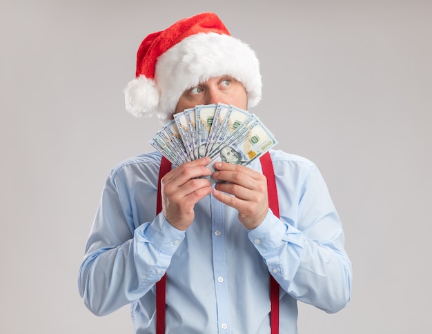 Jeune homme portant des bretelles noeud papillon in santa hat holding cash à côté surpris debout sur fond blanc