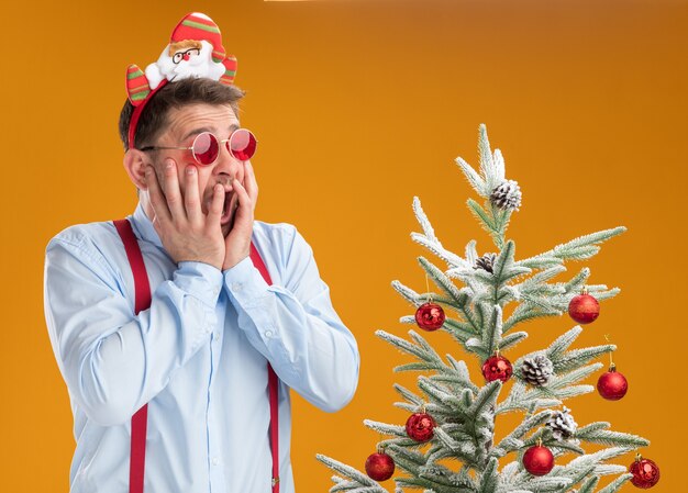 Jeune homme portant des bretelles noeud papillon en jante avec le père Noël et des lunettes rouges debout à côté de l'arbre de Noël à l'arbre en panique sur fond orange