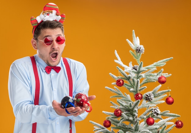 Jeune homme portant des bretelles noeud papillon en jante avec le père Noël et des lunettes rouges debout à côté de l'arbre de Noël à la confusion et étonné tenant des jouets pour arbre sur mur orange