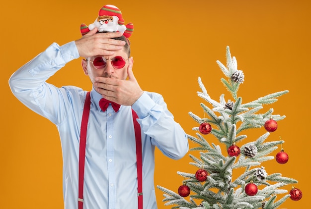 Jeune homme portant des bretelles noeud papillon en jante avec le père Noël et des lunettes rouges debout à côté de l'arbre de Noël avec la main sur son front couvrant la bouche avec l'autre main sur le mur orange