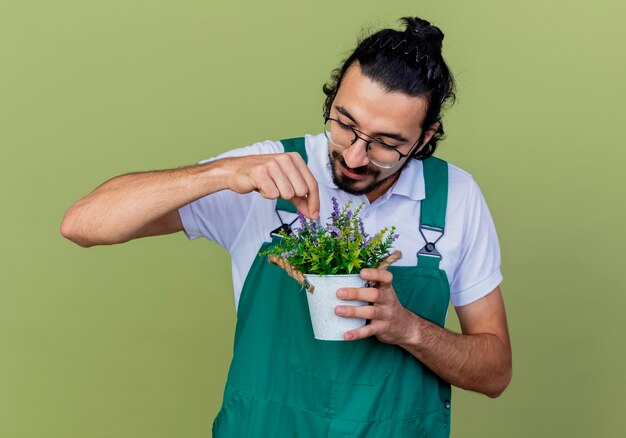 Jeune homme portant une combinaison de jardinier barbu tenant une plante en pot en souriant debout sur un mur vert clair