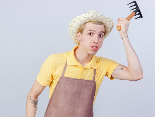 Jeune homme portant une combinaison de jardinier et un mini râteau balançant un chapeau étant confus