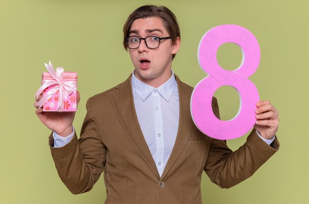 Jeune homme portant des lunettes tenant le numéro huit en carton et présent à l'avant heureux et surpris de la journée internationale de la femme debout sur le mur vert