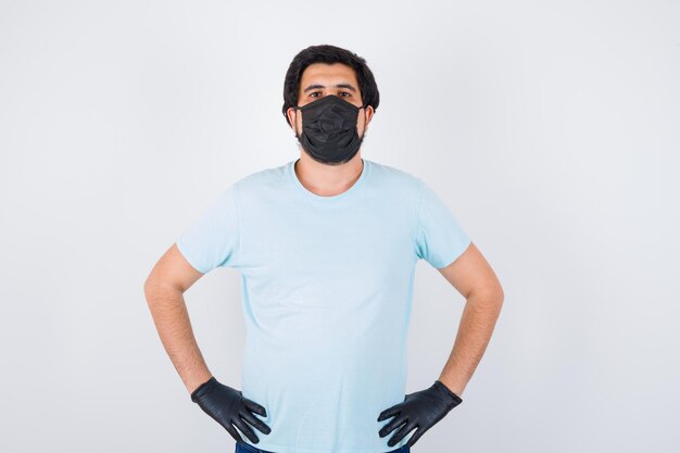 Jeune homme portant un masque médical et des gants tout en tenant les mains sur la taille en t-shirt et l'air confiant, vue de face.