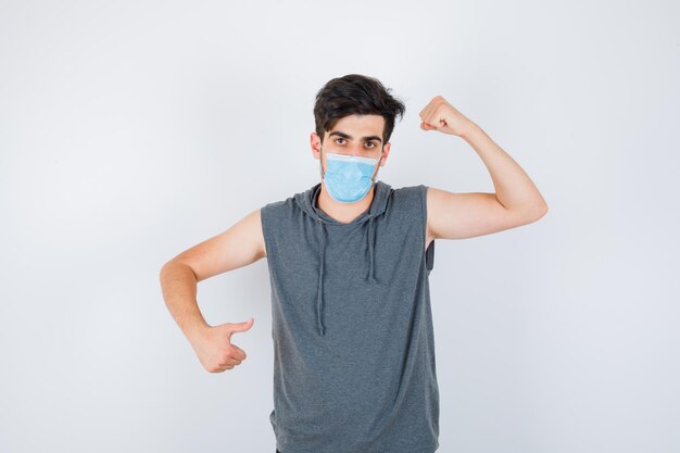 Jeune homme portant un masque tout en montrant le pouvoir et le geste du pouce en t-shirt gris et l'air sérieux