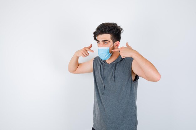 Jeune homme portant un masque tout en se pointant en t-shirt gris et l'air sérieux