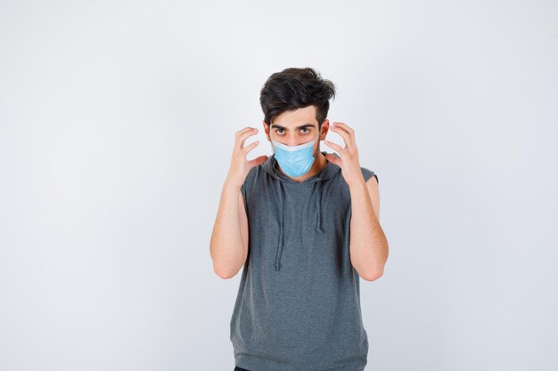 Jeune homme portant un masque tout en se tenant la main près du visage en t-shirt gris et l'air sérieux