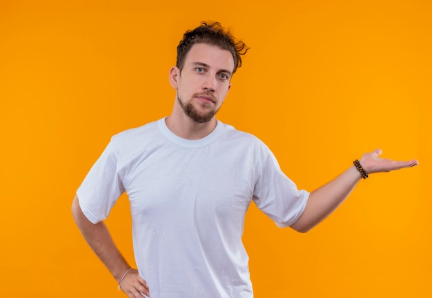 Jeune homme portant des points de t-shirt blanc à côté a mis sa main sur la hanche sur un mur orange isolé