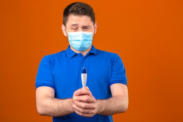 Jeune homme portant un polo bleu dans un masque de protection médicale à la recherche de thermomètre numérique à la main nerveux et inquiet sur mur orange isolé