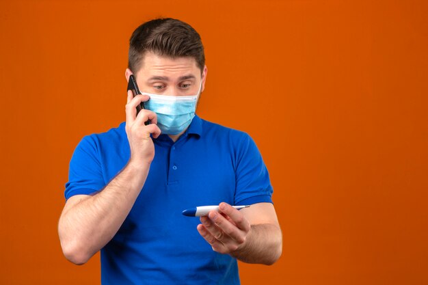 Jeune homme portant un polo bleu dans un masque de protection médicale à la recherche de thermomètre numérique à la main, parler au téléphone mobile à la nervosité et inquiet sur mur orange isolé