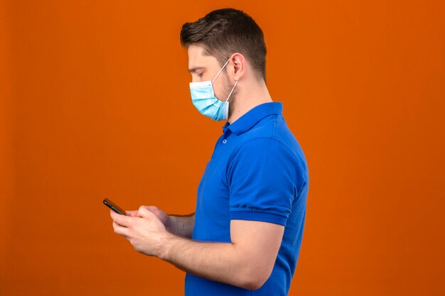 Jeune homme portant un polo bleu en masque de protection médicale debout sur le côté à la recherche de smartphone dans les mains sur un mur orange isolé