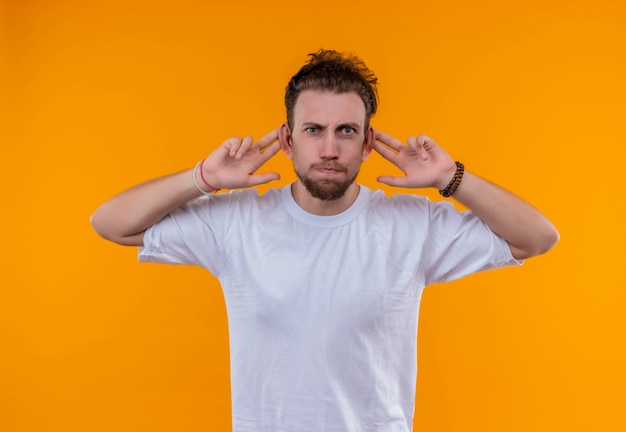 Jeune homme portant un t-shirt blanc a mis ses doigts sur les oreilles sur un mur orange isolé