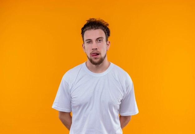 Jeune homme portant un t-shirt blanc a mis ses mains sur le dos et montrant la langue sur un mur orange isolé