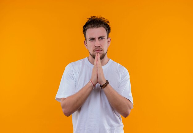 Jeune homme portant un t-shirt blanc montrant le geste de prier sur un mur orange isolé