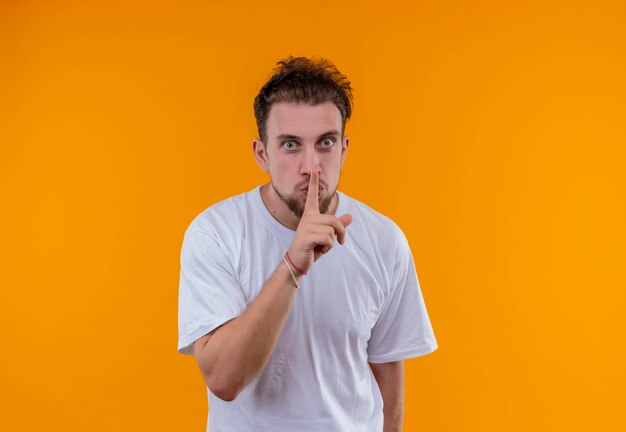 Jeune homme portant un t-shirt blanc montrant le geste de silence sur un mur orange isolé