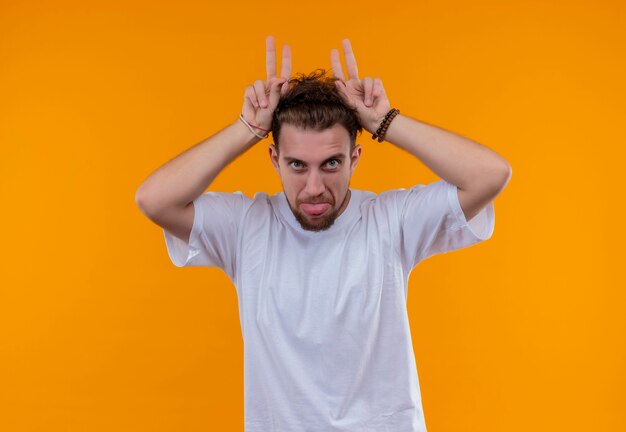 Jeune homme portant un t-shirt blanc montrant la langue et les oreilles de lapin geste sur mur orange isolé