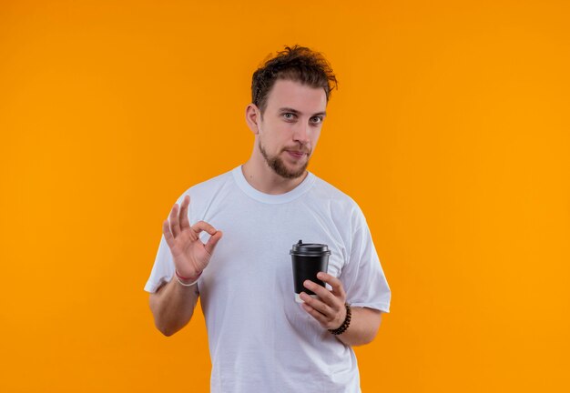 Jeune homme portant un t-shirt blanc tenant une tasse de café montrant le geste okey sur mur orange isolé