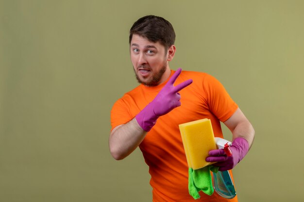 Jeune homme portant un t-shirt orange et des gants en caoutchouc tenant des outils de nettoyage confiants et heureux montrant le numéro deux ou signe de la victoire debout sur le mur vert