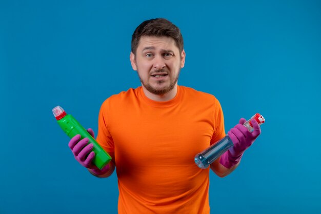 Jeune homme portant un t-shirt orange et des gants en caoutchouc tenant des produits de nettoyage b