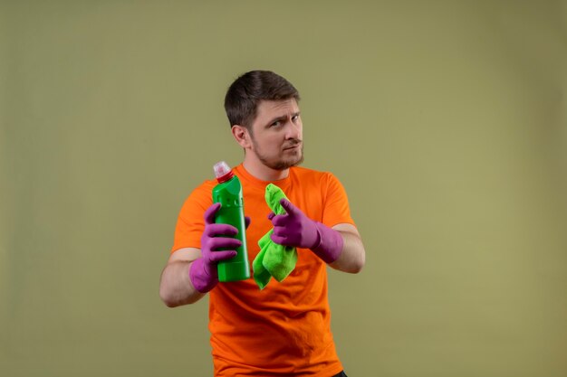 Jeune homme portant un t-shirt orange et des gants en caoutchouc tenant des produits de nettoyage à la confiance