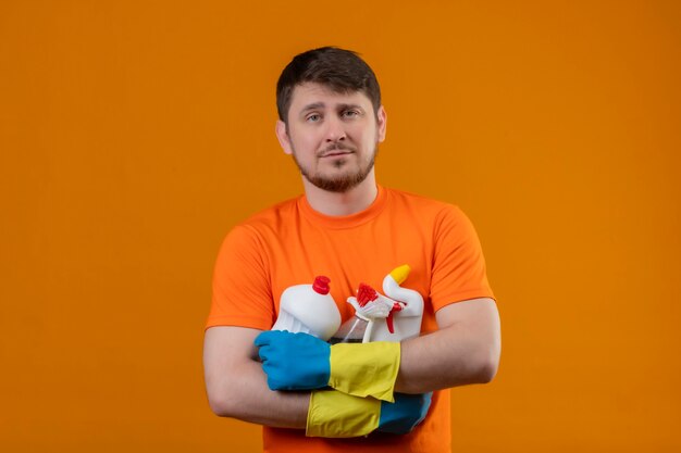 Jeune homme portant un t-shirt orange et des gants en caoutchouc tenant des produits de nettoyage avec une expression confiante sérieuse