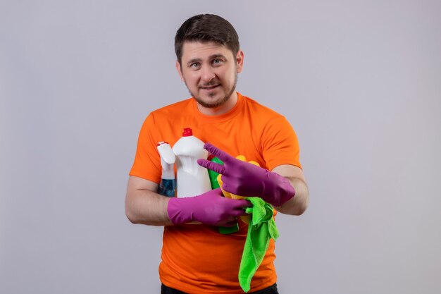 Photo gratuite jeune homme portant un t-shirt orange et des gants en caoutchouc tenant des produits de nettoyage montrant le numéro deux