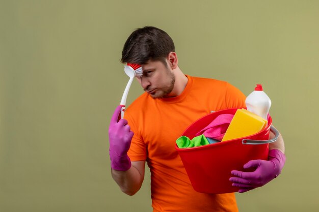 Jeune homme portant un t-shirt orange et des gants en caoutchouc tenant un seau avec des outils de nettoyage et une brosse à récurer