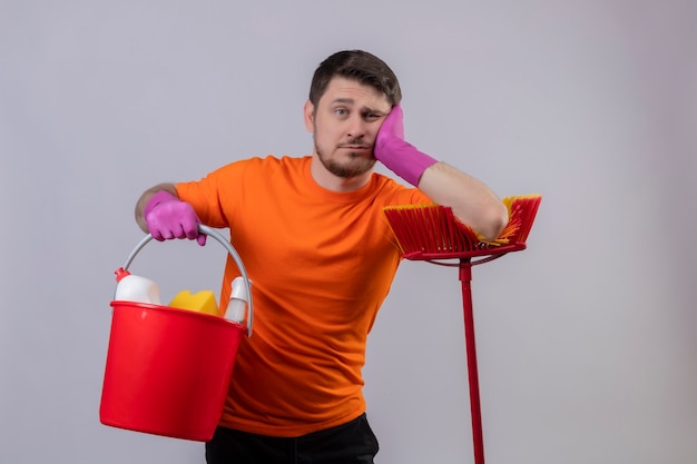 Photo gratuite jeune homme portant un t-shirt orange et des gants en caoutchouc tenant un seau avec des outils de nettoyage et une vadrouille à la fatigue et s'ennuie debout sur un mur blanc