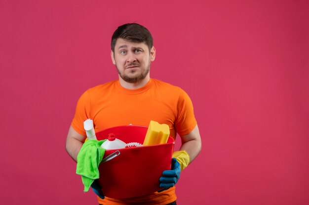 Jeune homme portant un t-shirt orange et des gants en caoutchouc tenant un seau avec des outils de nettoyage