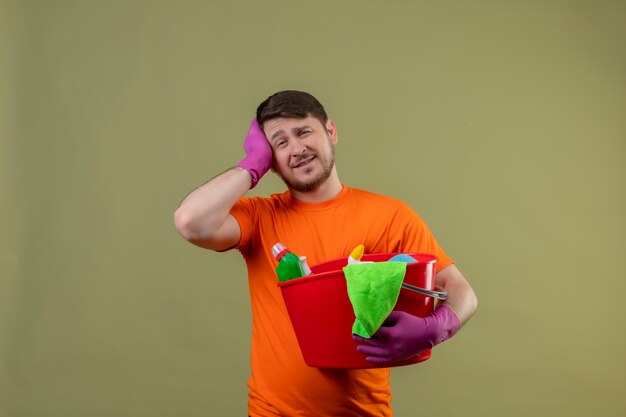 Jeune homme portant un t-shirt orange et des gants en caoutchouc tenant un seau avec des outils de nettoyage