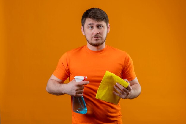 Jeune homme portant un t-shirt orange et des gants en caoutchouc tenant un spray de nettoyage et un tapis mécontent d'une expression triste